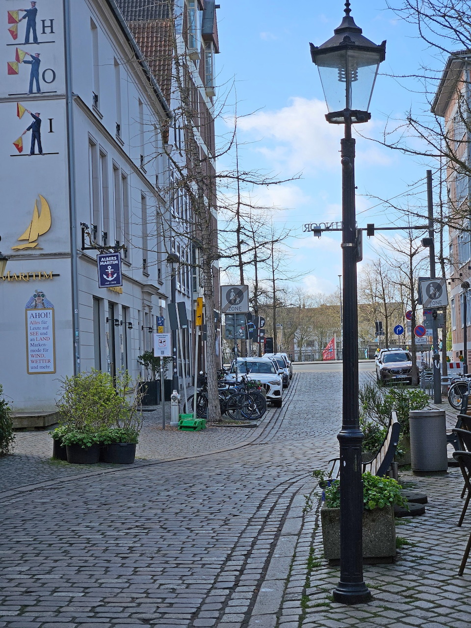 Cobblestone street in Bremen with historic buildings, parked cars, bare trees, and a street lamp