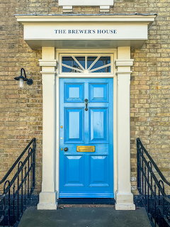 Bright blue paneled front door with brass letterbox and knocker beneath a sign reading The Brewers House