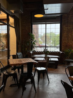 Coffee shop interior with wooden tables, metal chairs, leather couch, plants, and large window with street view