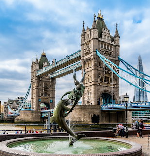 Bronze fountain statue of girl with dolphin in front of Tower Bridge, London, with blue sky and clouds
