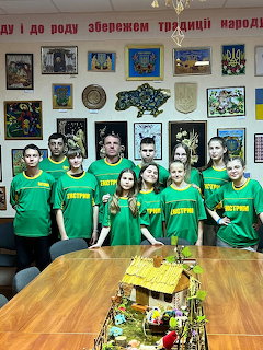 Group of young people in green t-shirts posing in front of a wall with framed artwork and a miniature house model