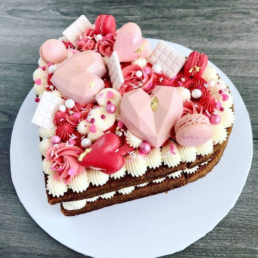 Heart-shaped chocolate cake decorated with pink macarons, strawberries, meringues, white chocolate, and pearl sprinkles