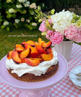 Rustic cake with whipped cream and sliced peaches on a pink stand beside flowers outdoors