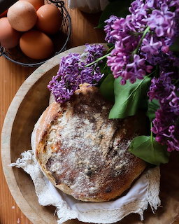 Round sourdough loaf on a wooden tray with purple lilacs and a bowl of brown eggs