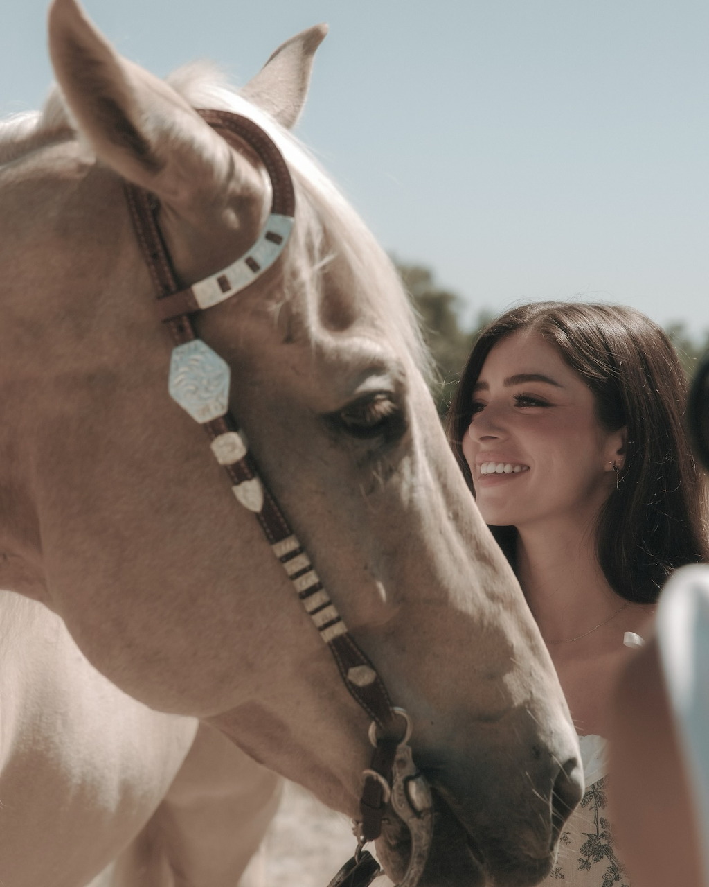 Smiling young woman next to a pale horse with a decorative bridle outdoors
