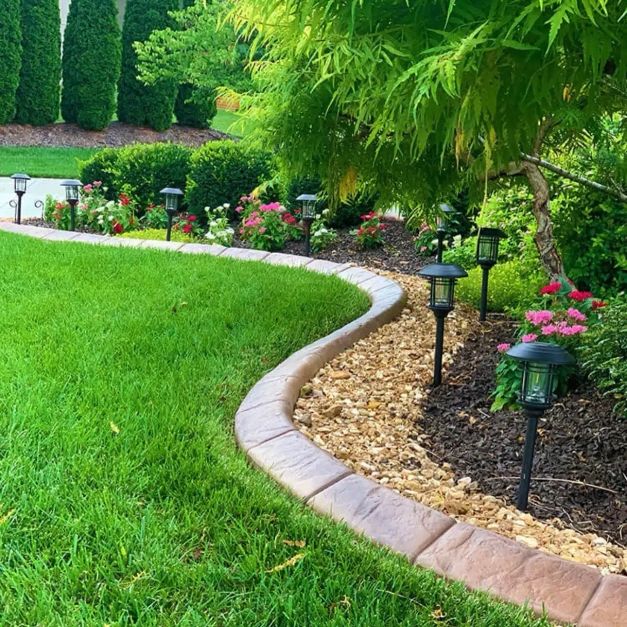 Curved stone-edged garden path with mulch, green lawn, flowers, and solar lights surrounded by lush greenery