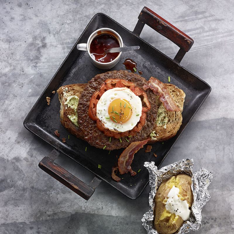 Grilled steak with fried egg on black griddle pan, coffee cup and baked potato on marble surface