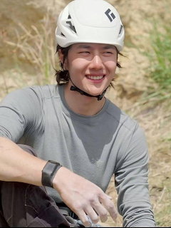 Smiling young man in white climbing helmet and gray shirt sitting outdoors on dry ground