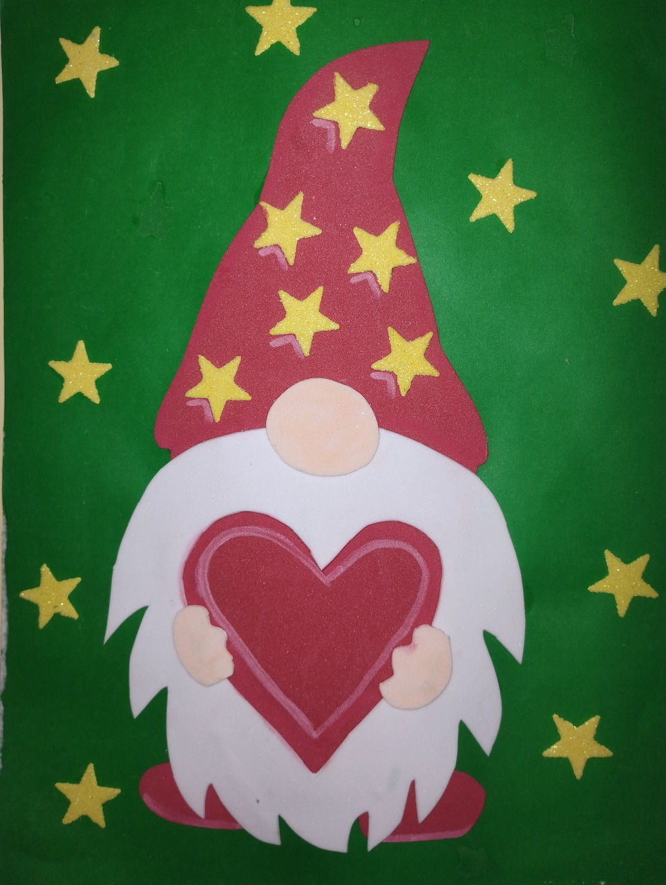 Paper craft gnome with red starred hat holding a red heart on green background with gold stars