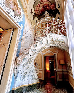 Ornate white baroque staircase with roco decorations, ceiling frescoes, and wrought iron railing in a palace interior
