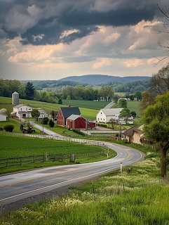 Winding country road passing red barn and farmhouses amid green fields under dramatic cloudy sky with mountains