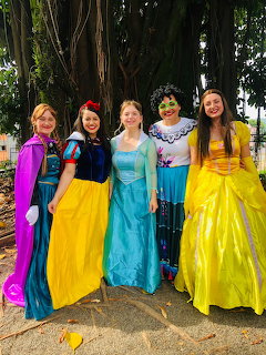 Five women in princess costumes standing together outdoors in colorful gowns
