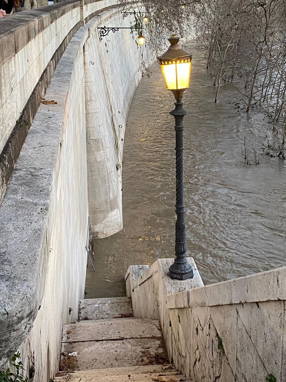 Lit street lamp on stone steps leading down to a flooded river beside a curved stone wall