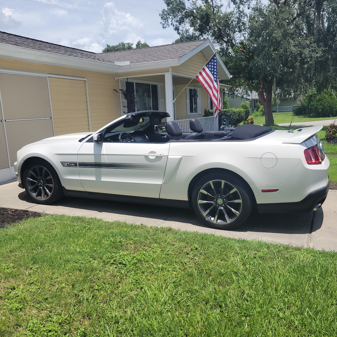 White Ford Mustang convertible parked in a driveway beside a house with an American flag