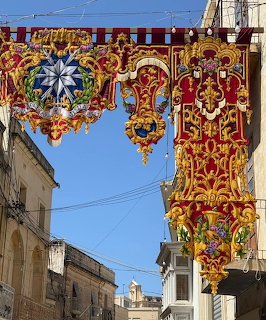 Elaborate red and gold decorative banners with baroque designs suspended over a Malta street