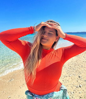 Woman in orange swim top poses at beach with blue ocean and sky behind her