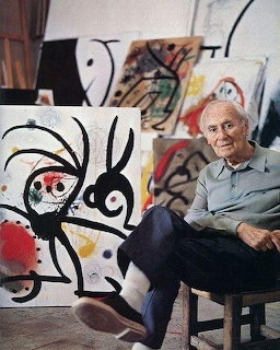 Elderly man in blue shirt sitting in art studio surrounded by colorful abstract paintings