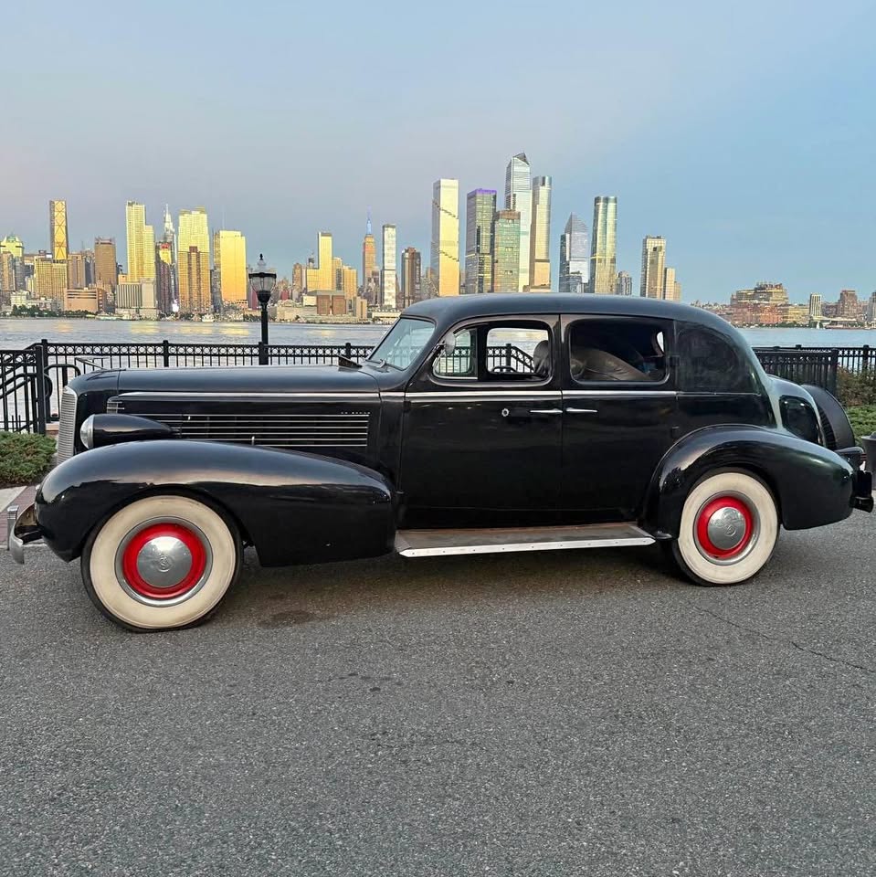 Black 1937 LaSalle sedan parked by the water with a city skyline behind it