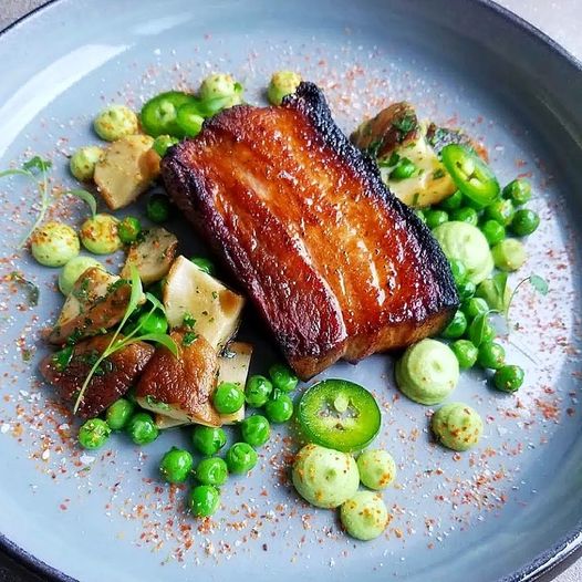 Crispy pork belly plated with peas, jalapeño slices, and small green vegetables on a gray plate