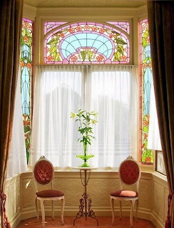Bay window with ornate Art Nouveau stained glass panels, white curtains, two vintage chairs and a potted plant