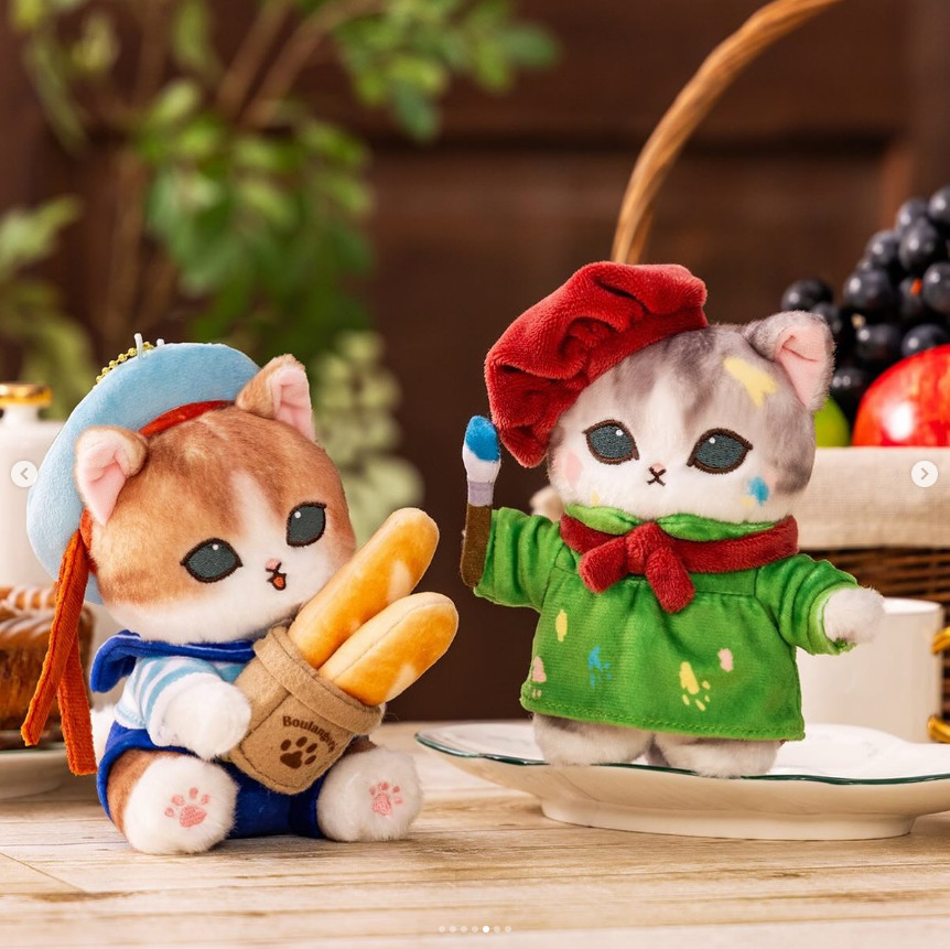Two Mofusand cat plush toys in colorful clothes on a table, one holding bread and one waving