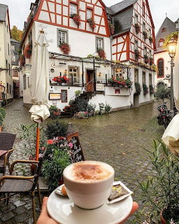 Hand holding a latte cup in front of half-timbered buildings on a cobblestone street in Beilstein, Germany
