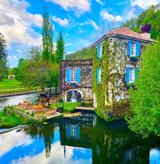 Stone cottage with blue shutters beside a calm river, reflected in the water
