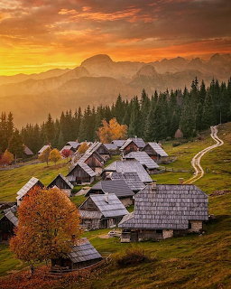 Wooden village houses on hillside with forest, mountains, and vibrant sunset
