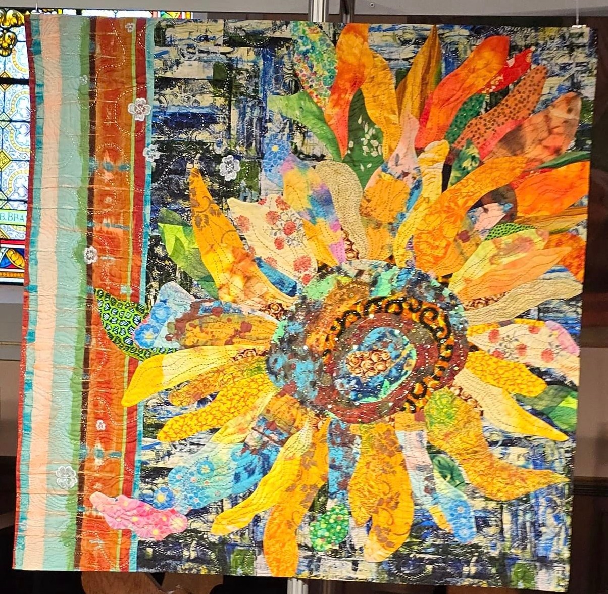 Colorful fabric collage artwork depicting a large sunflower with orange and yellow petals against a blue patchwork background
