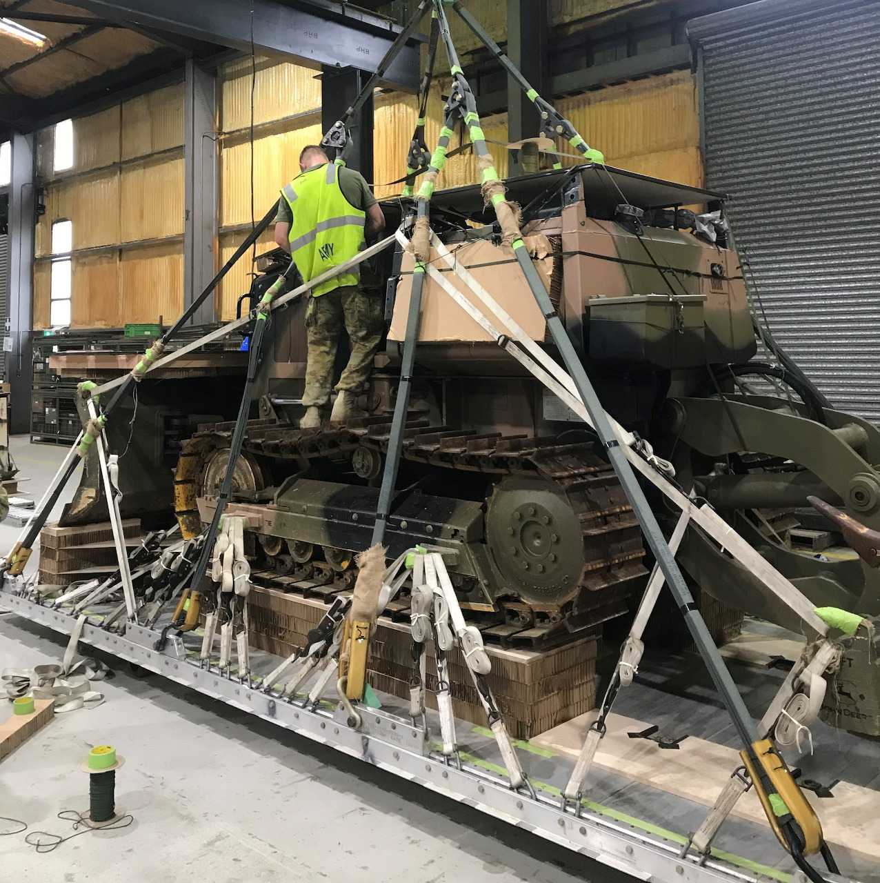 Tracked military vehicle secured with straps in a warehouse, with two workers standing on top