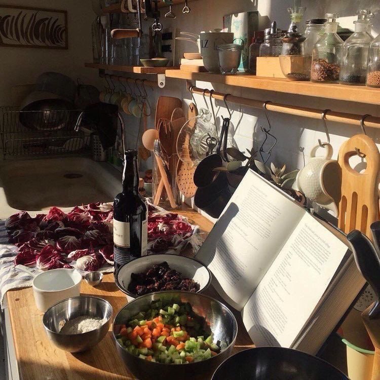 Rustic kitchen counter with open cookbook, pan of colorful vegetables, wooden utensils, and shelves with glass jars