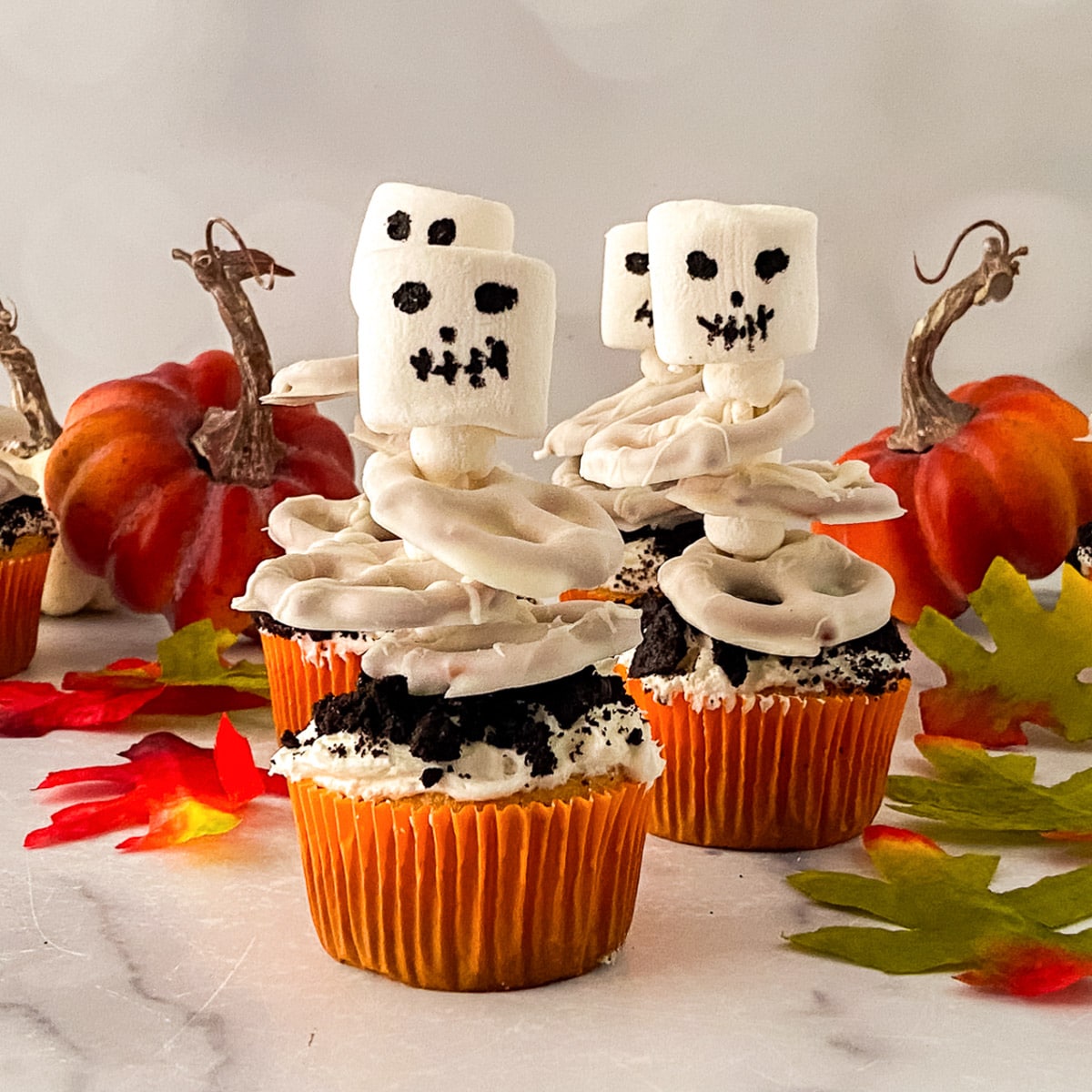 Three Halloween cupcakes topped with ghost marshmallows, with mini pumpkins and fall leaves behind them