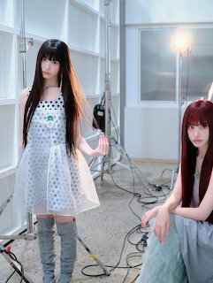 Two young women posing in a bright studio with long hair, white dresses, and lighting equipment