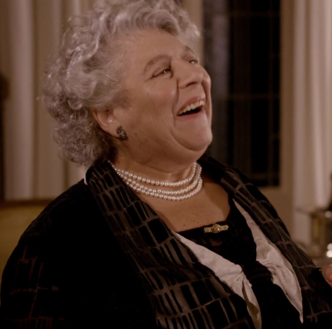 Elderly woman laughing indoors, wearing pearl necklaces and a dark outfit