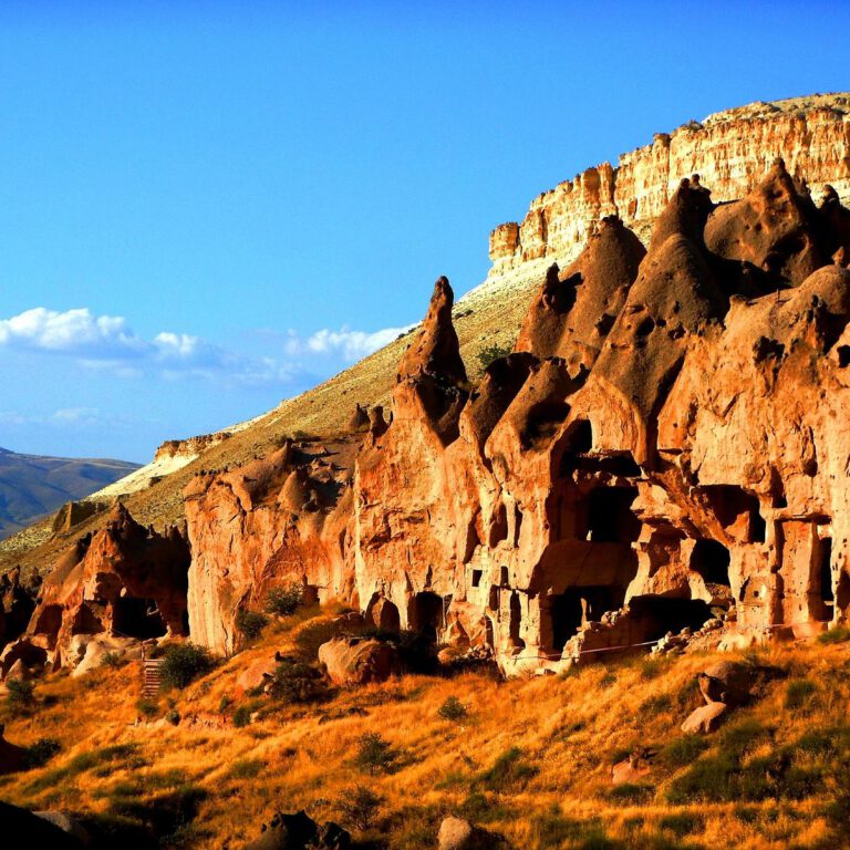 Ancient cave dwellings carved into rocky cliffs on a dry hillside beneath a blue sky