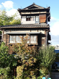 Old weathered two-story Japanese wooden house with dark siding and traditional tiled roof surrounded by greenery