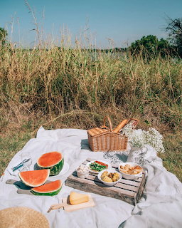 Picnic blanket with wicker basket, watermelon, bread, cheese, and white flowers in a grassy field
