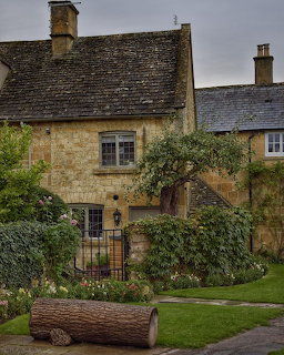 Stone cottage with climbing plants, green lawn, and log border under cloudy sky