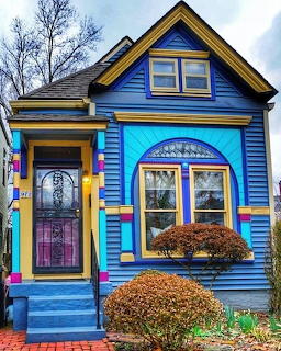Bright blue Victorian house with yellow and pink trim, ornate door and windows, front steps and landscaped shrubs
