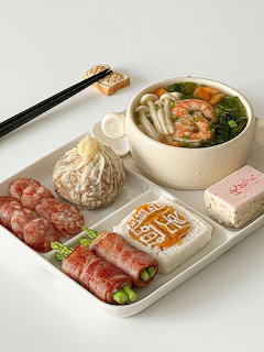 White tray with Korean food including seafood soup, tofu, rolled meat, sausages, dumplings, and chopsticks