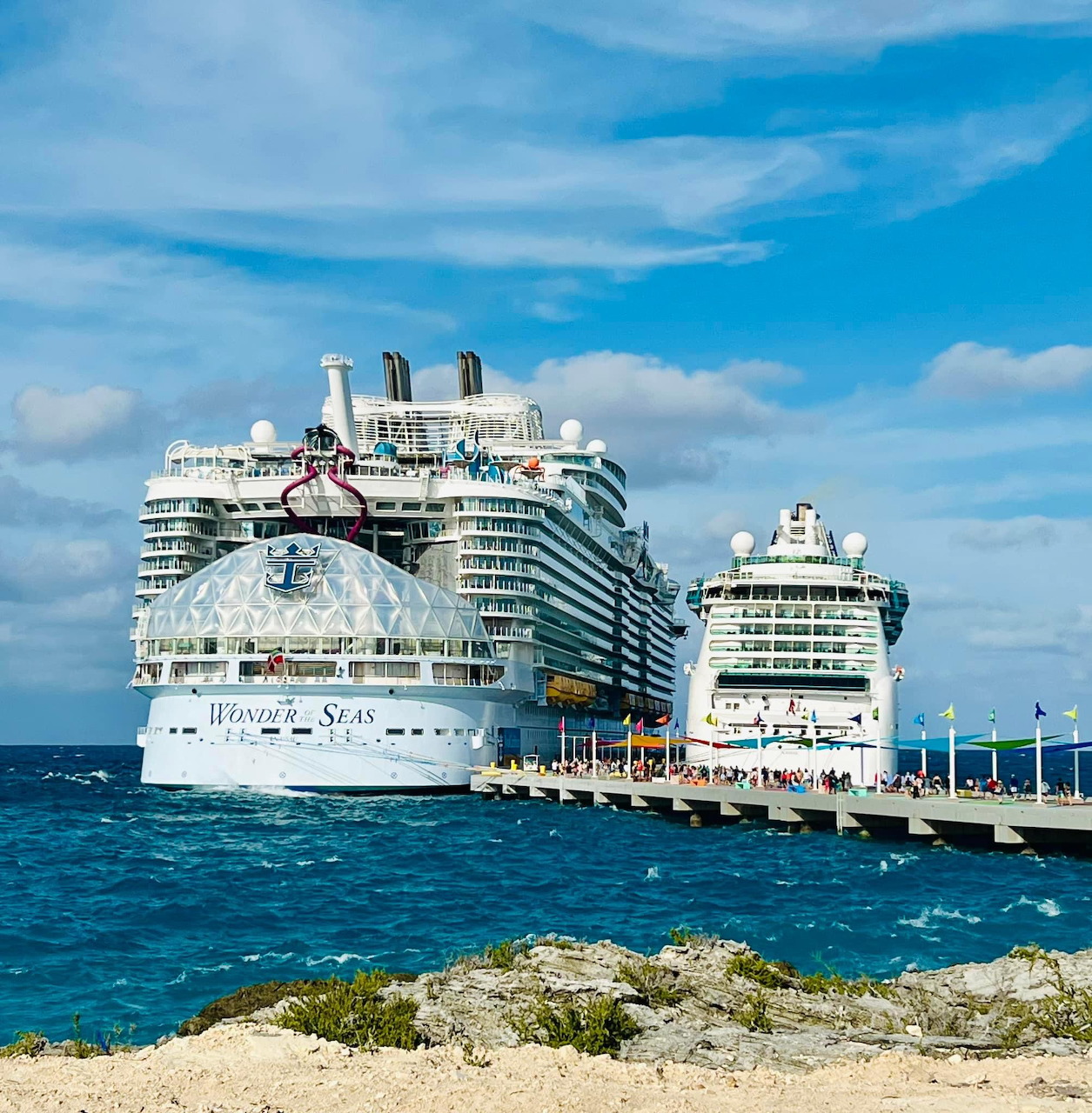 Two white cruise ships docked at a pier on blue ocean water beneath a cloudy sky