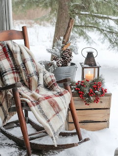 Wooden rocking chair with plaid blanket in snow beside a lantern and Christmas decorations