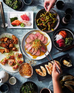 Overhead shot of Japanese dishes including sashimi, grilled meat, vegetables, and sushi on a table