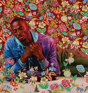 Man in blue denim and purple jacket seated against colorful floral patterned background