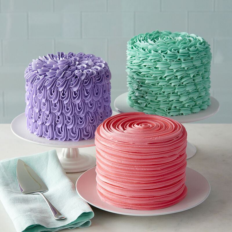 Three colorful frosted cakes with different piped icing patterns on white cake stands