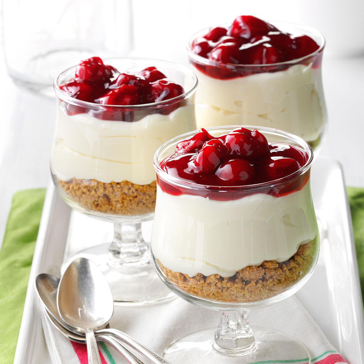 Three cherry cheesecake parfaits in glass cups with layered graham crust, cream filling, and cherry topping