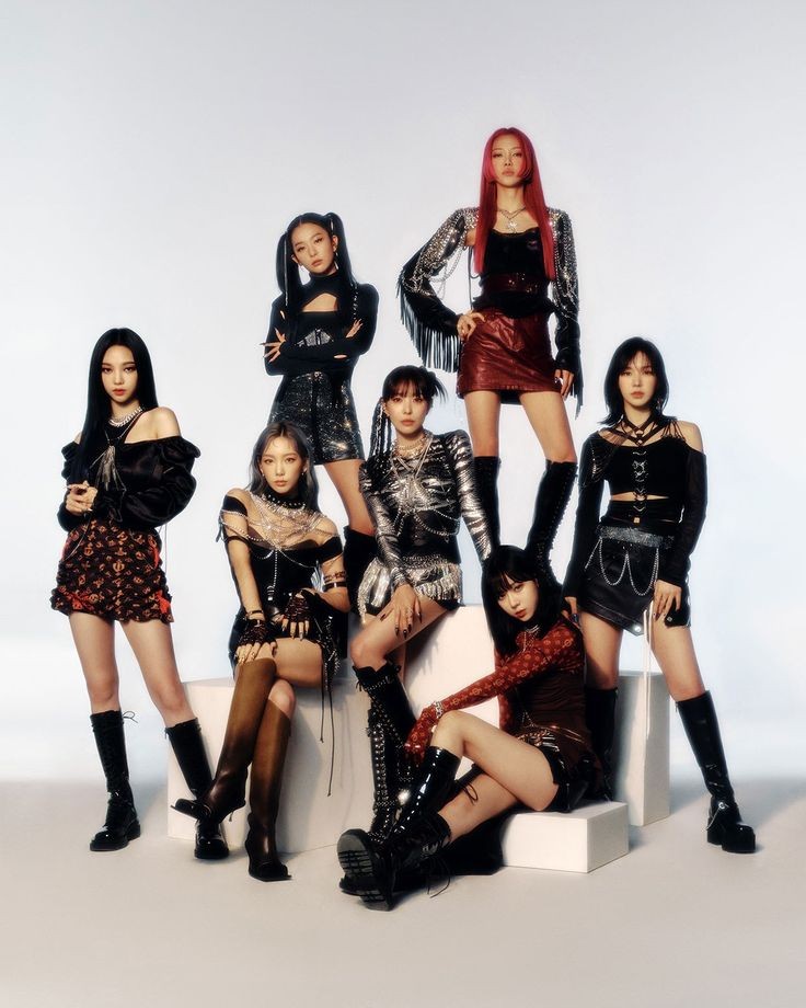 Seven women in coordinated black outfits with red accents posing as a K-pop group