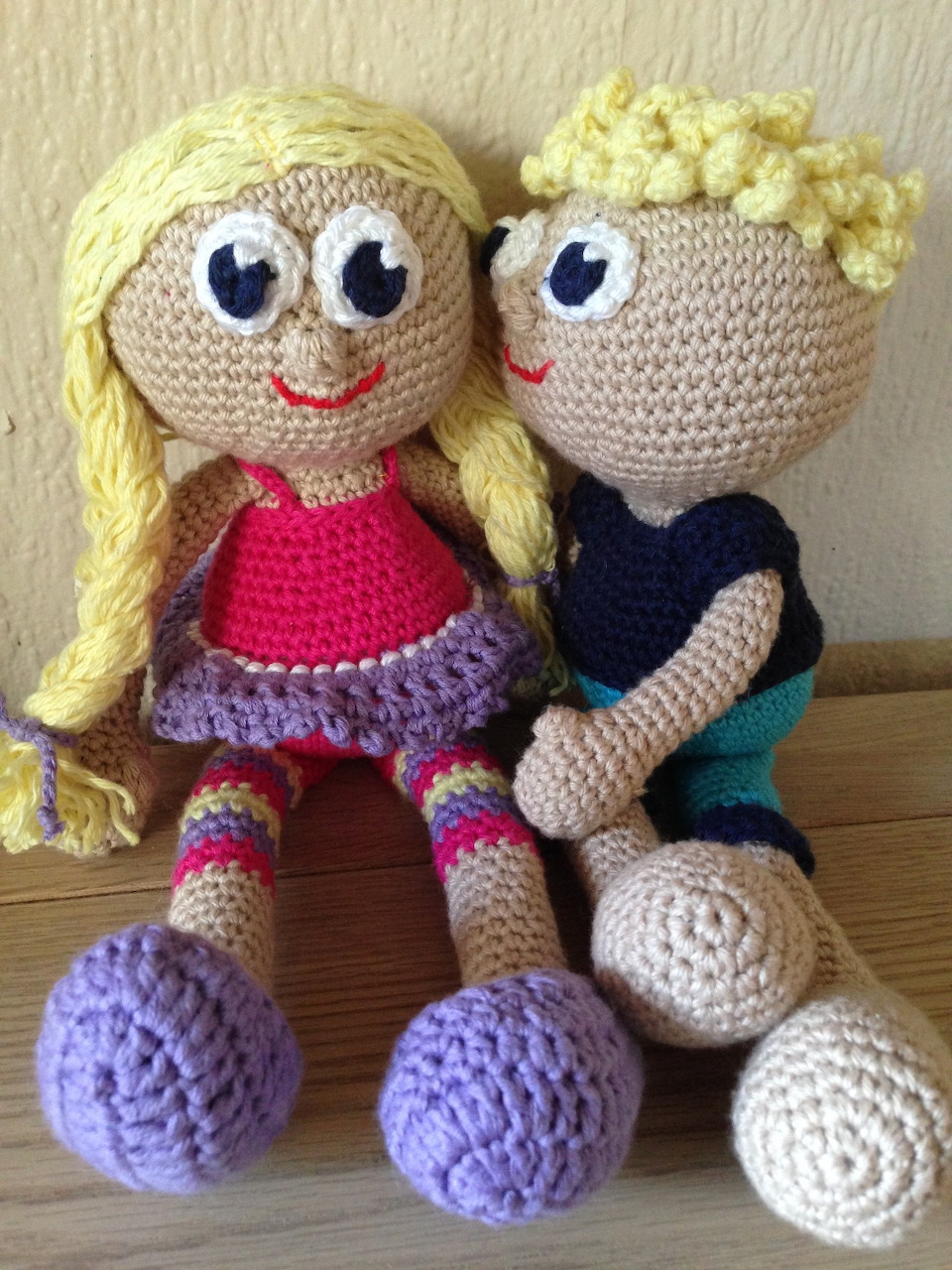 Two crocheted amigurumi dolls with blonde yarn hair sitting side by side on wood