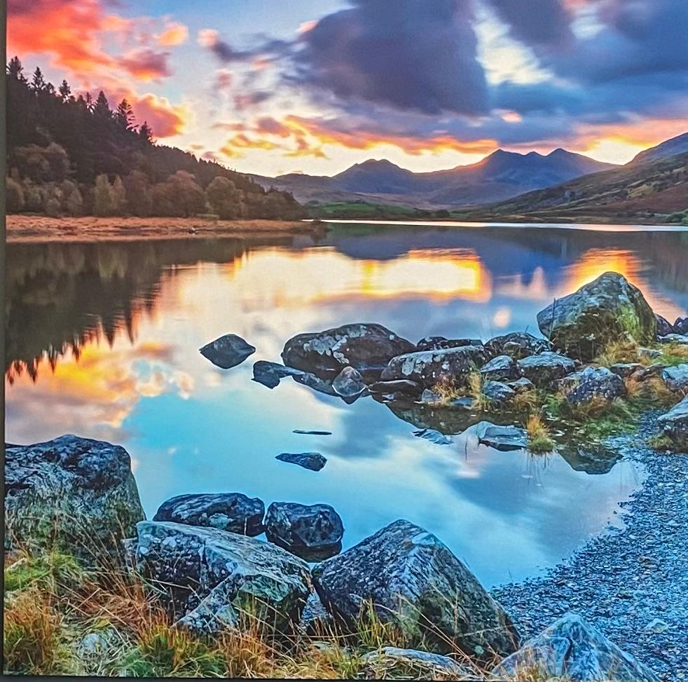Mountain lake at sunset with vibrant orange and blue sky reflecting on calm water, rocks scattered in foreground