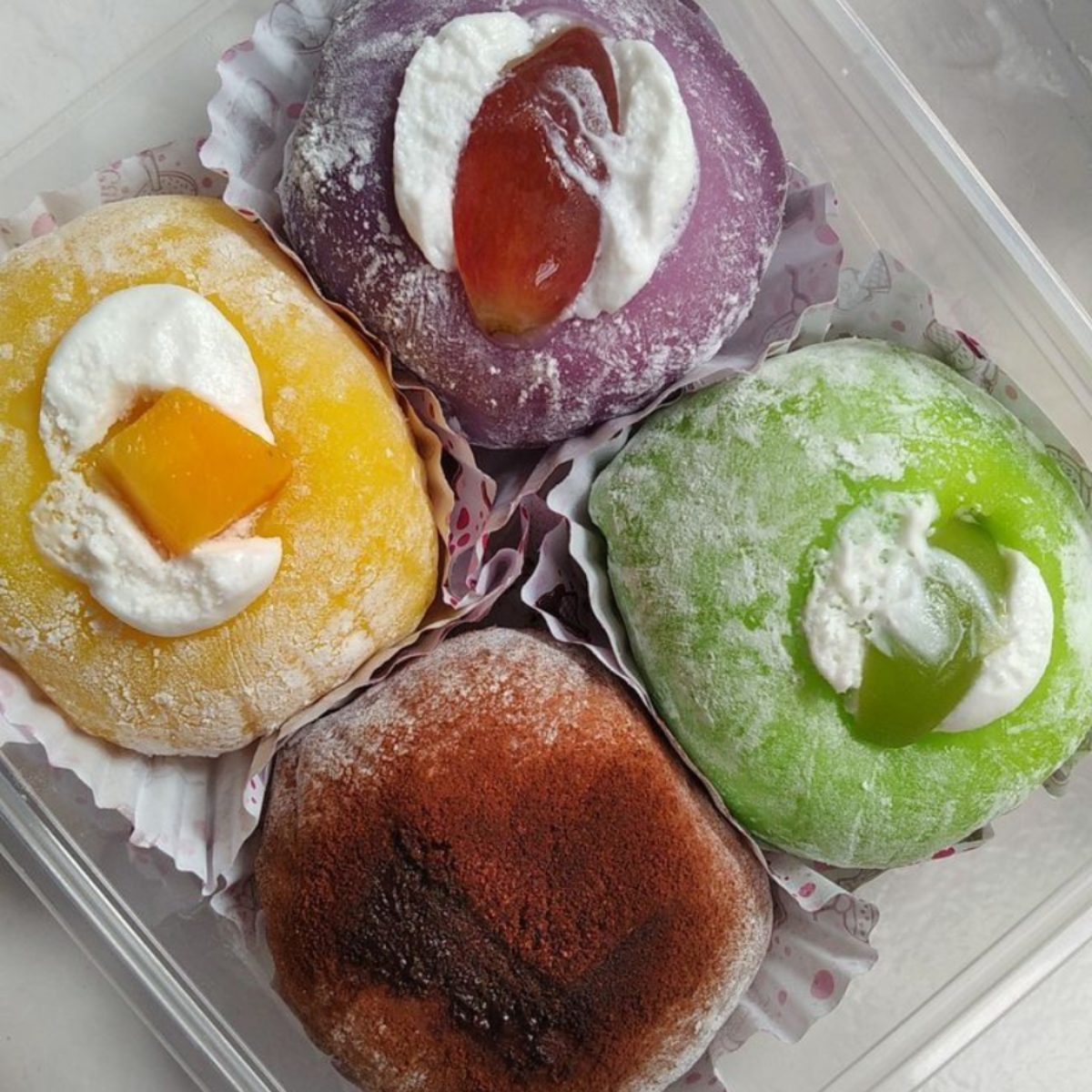 Four colorful mochi balls with fruit centers in yellow, purple, green and brown, dusted with powdered sugar